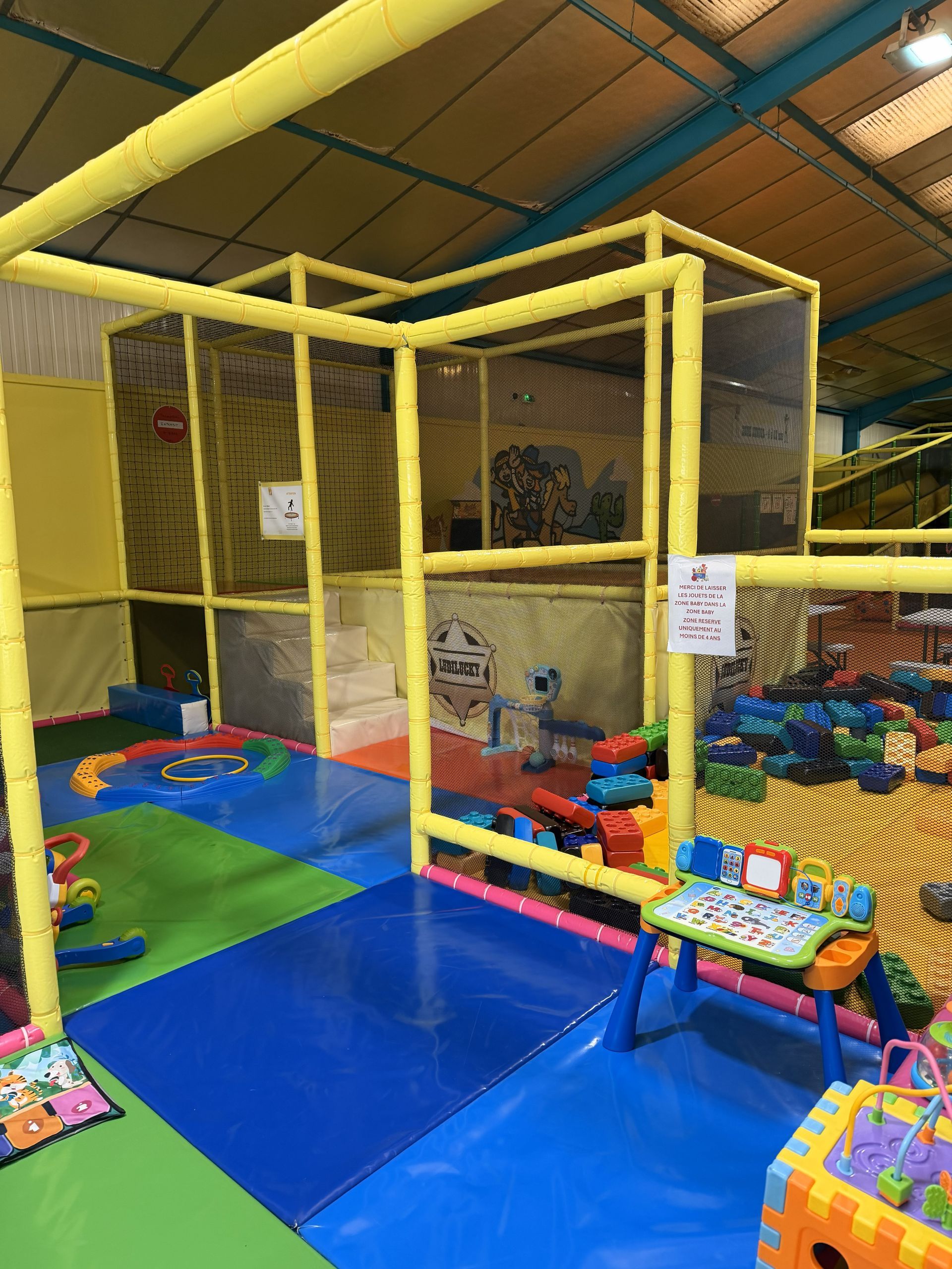 Aire de jeux intérieure avec structure de jeu jaune à plusieurs niveaux et tapis colorés.