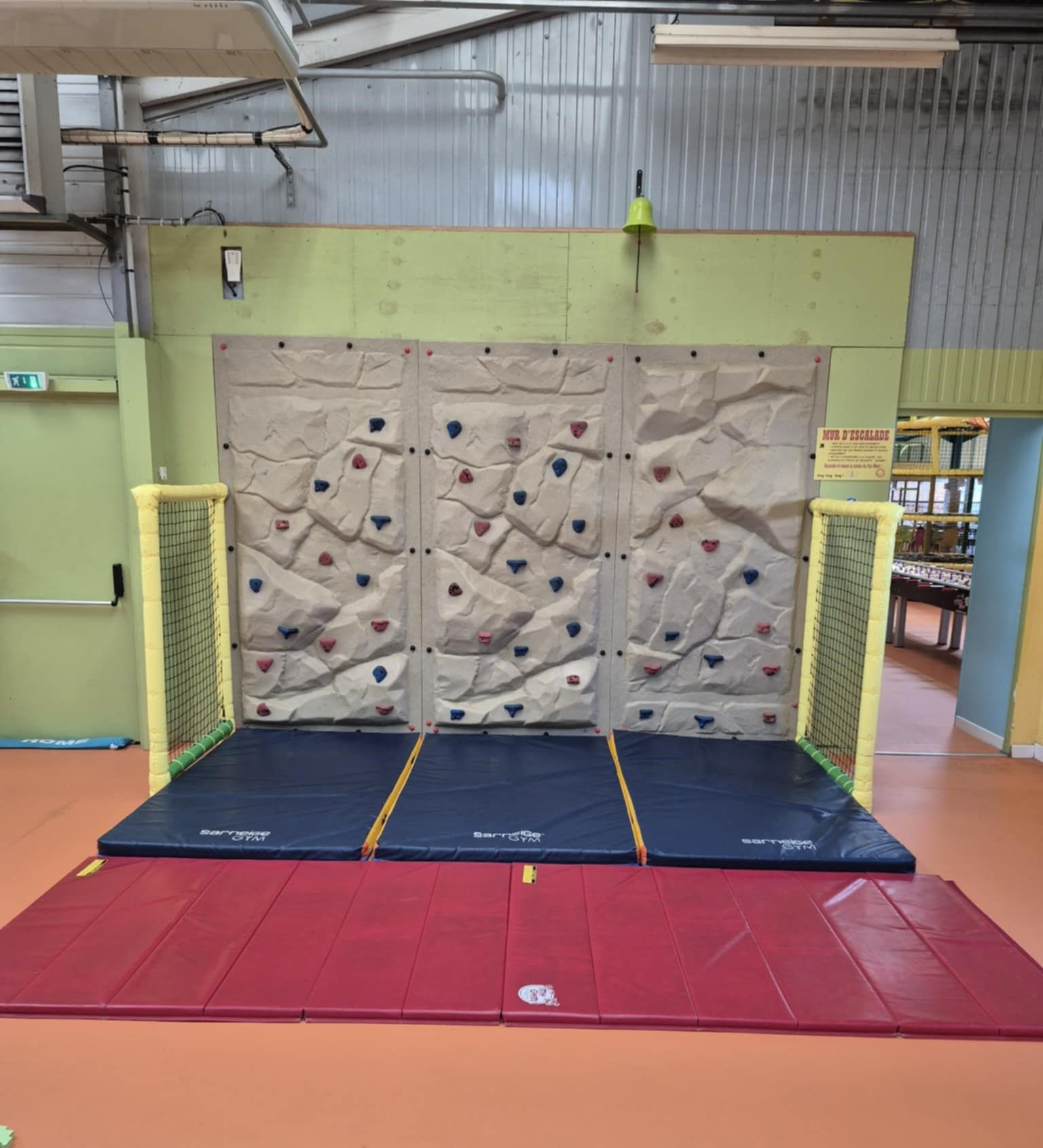 Mur d'escalade intérieur avec tapis de sécurité et barrières de protection jaunes.