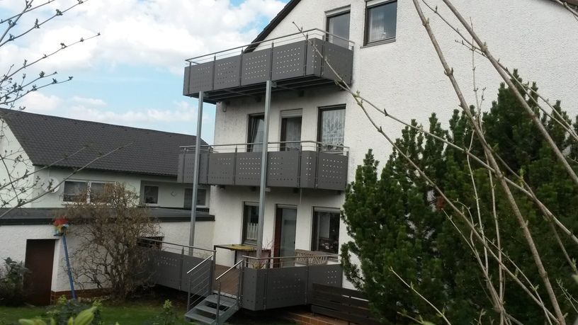Ein weißes zweistöckiges Haus mit grauen Balkonen, die durch Treppen verbunden sind. Grüne Büsche und blauer Himmel im Hintergrund.