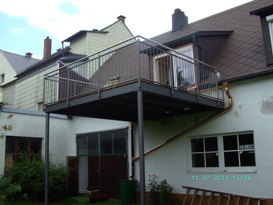 Ein dunkelgrauer Balkon an einem weißen Gebäude. Metallgeländer, Metallpfosten. Dahinter ein weiteres Gebäude.