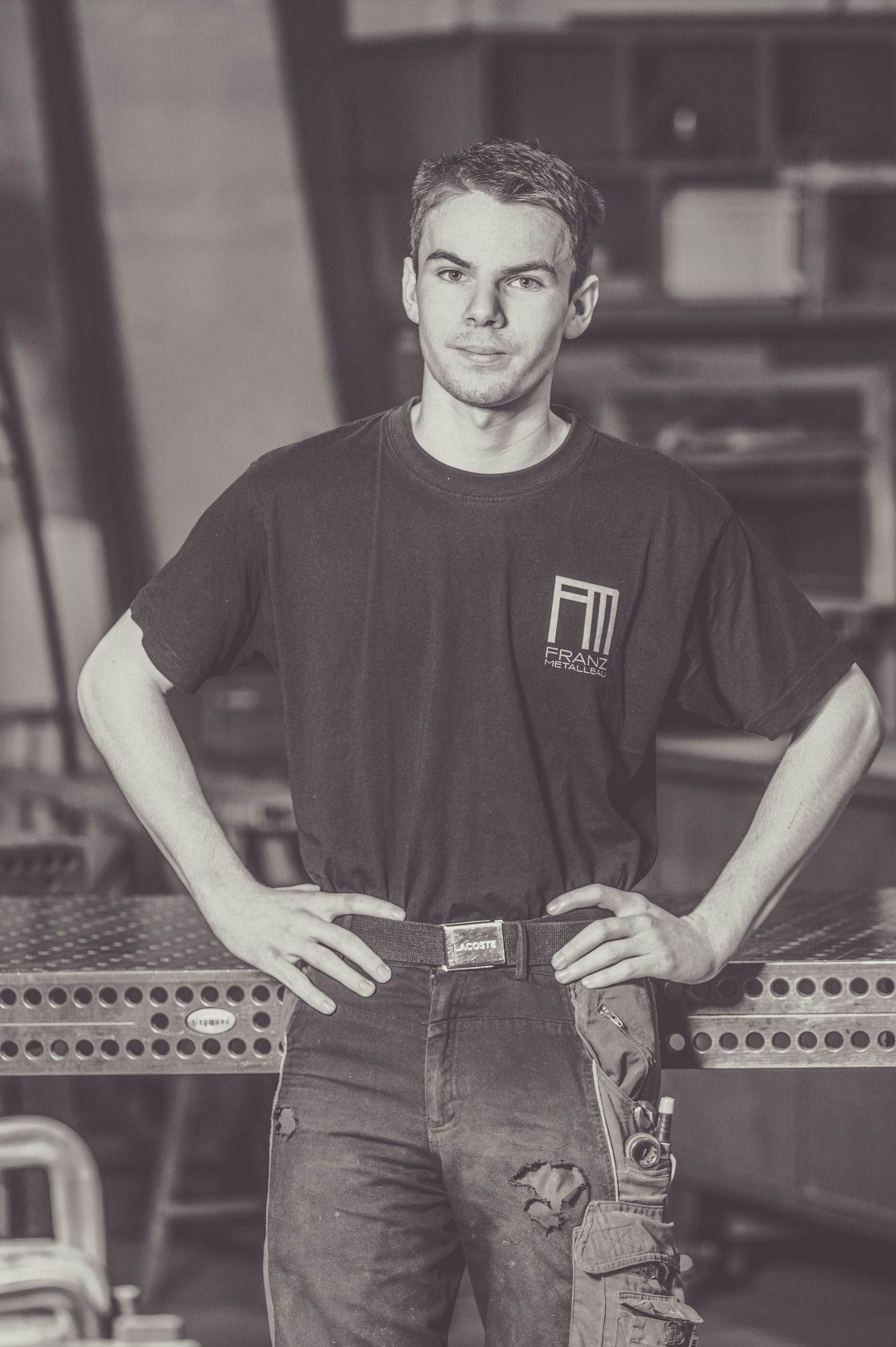 Mann posiert in einem Geschäft mit den Händen in den Hüften. Er trägt ein schwarzes T-Shirt und Jeans in einer Werkstattumgebung.