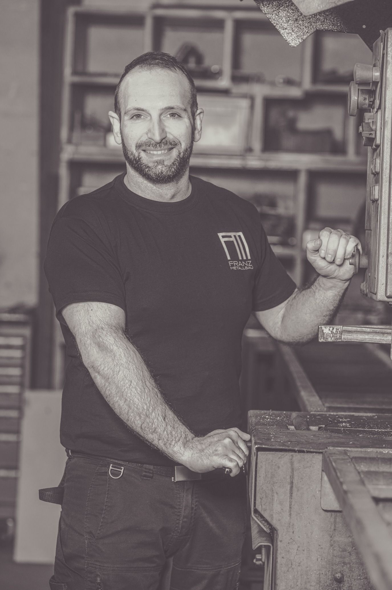 Mann in einer Werkstatt lächelt, trägt ein schwarzes Hemd und hält eine Holzkonstruktion.