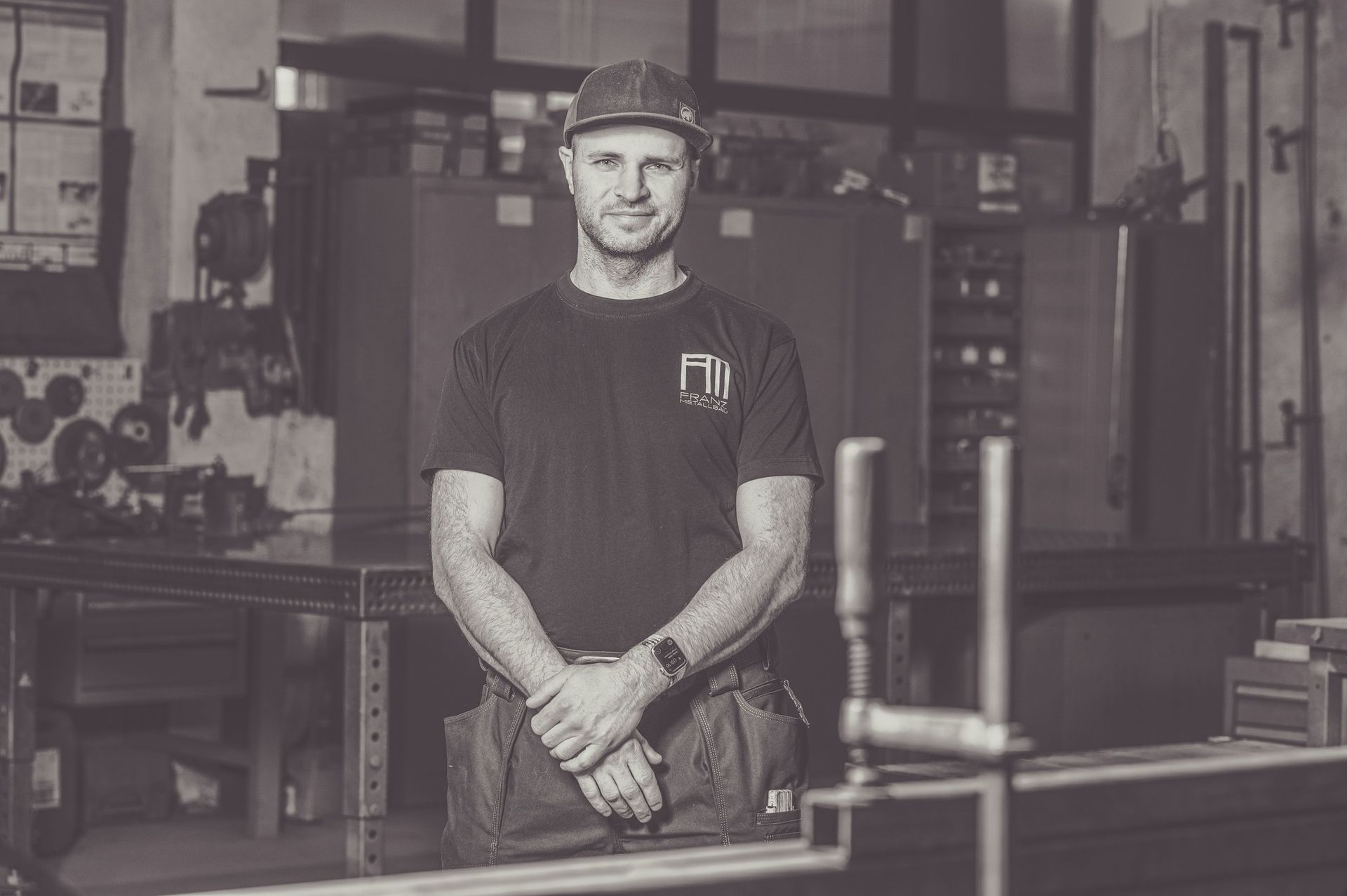 Mann in der Werkstatt mit Hut, verschränkten Armen, lächelnd. Industrielle Umgebung mit sichtbaren Maschinen.