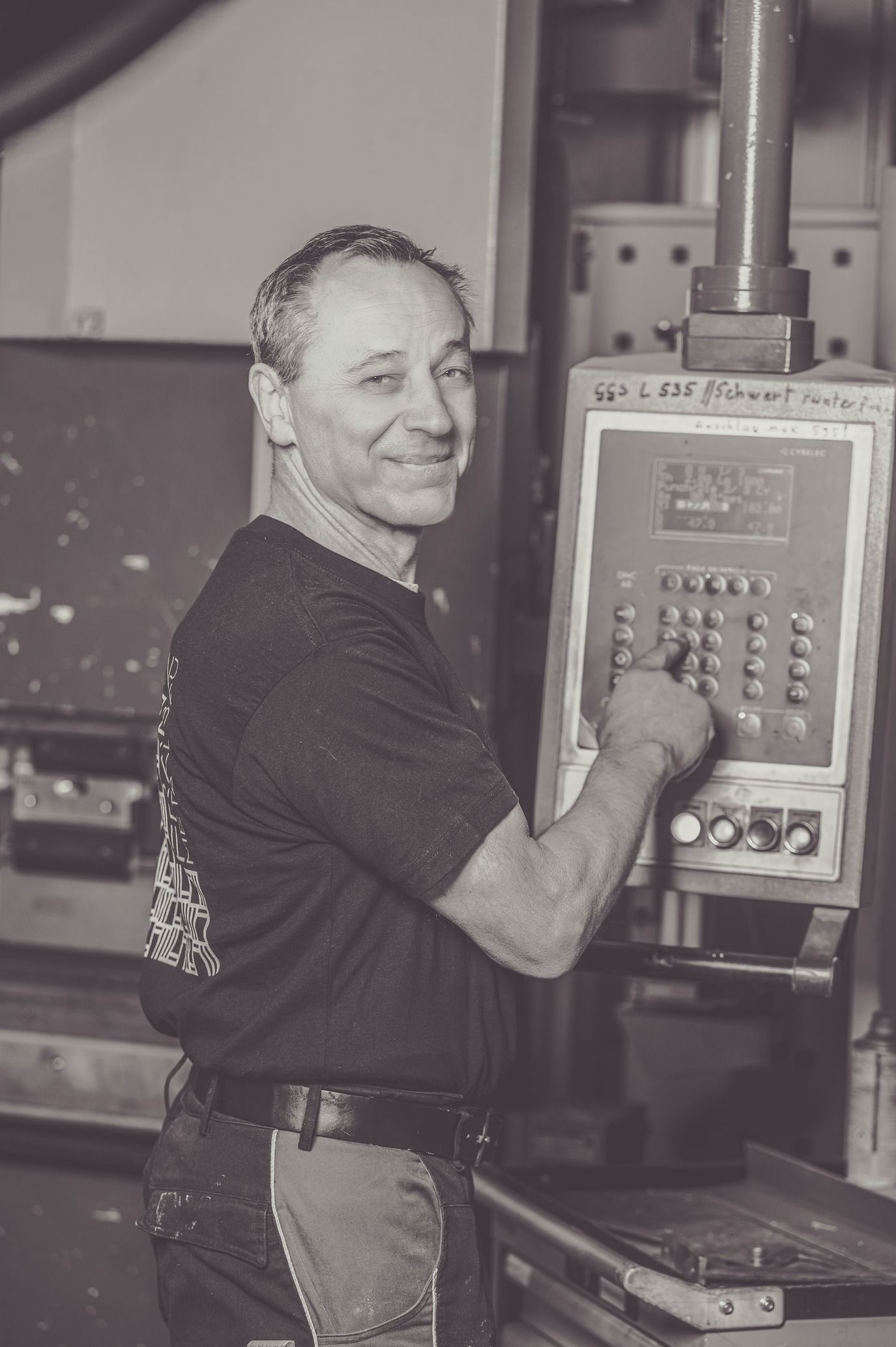 Mann in Werkstatt bedient Maschinen, lächelnd, schwarzweiß.