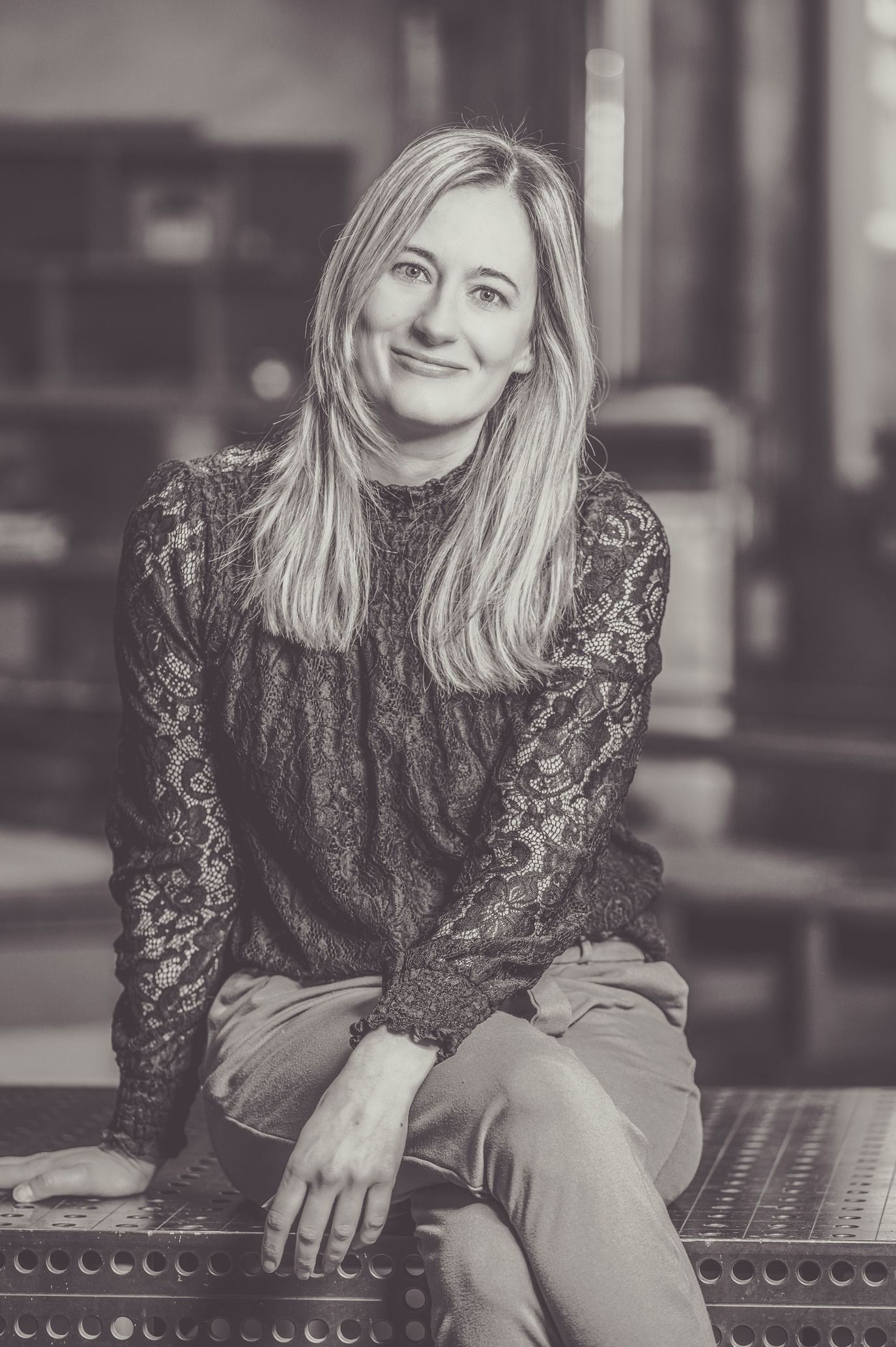 Frau mit langen blonden Haaren lächelt und sitzt mit gekreuzten Beinen. Sie trägt ein Spitzenhemd und eine helle Hose.