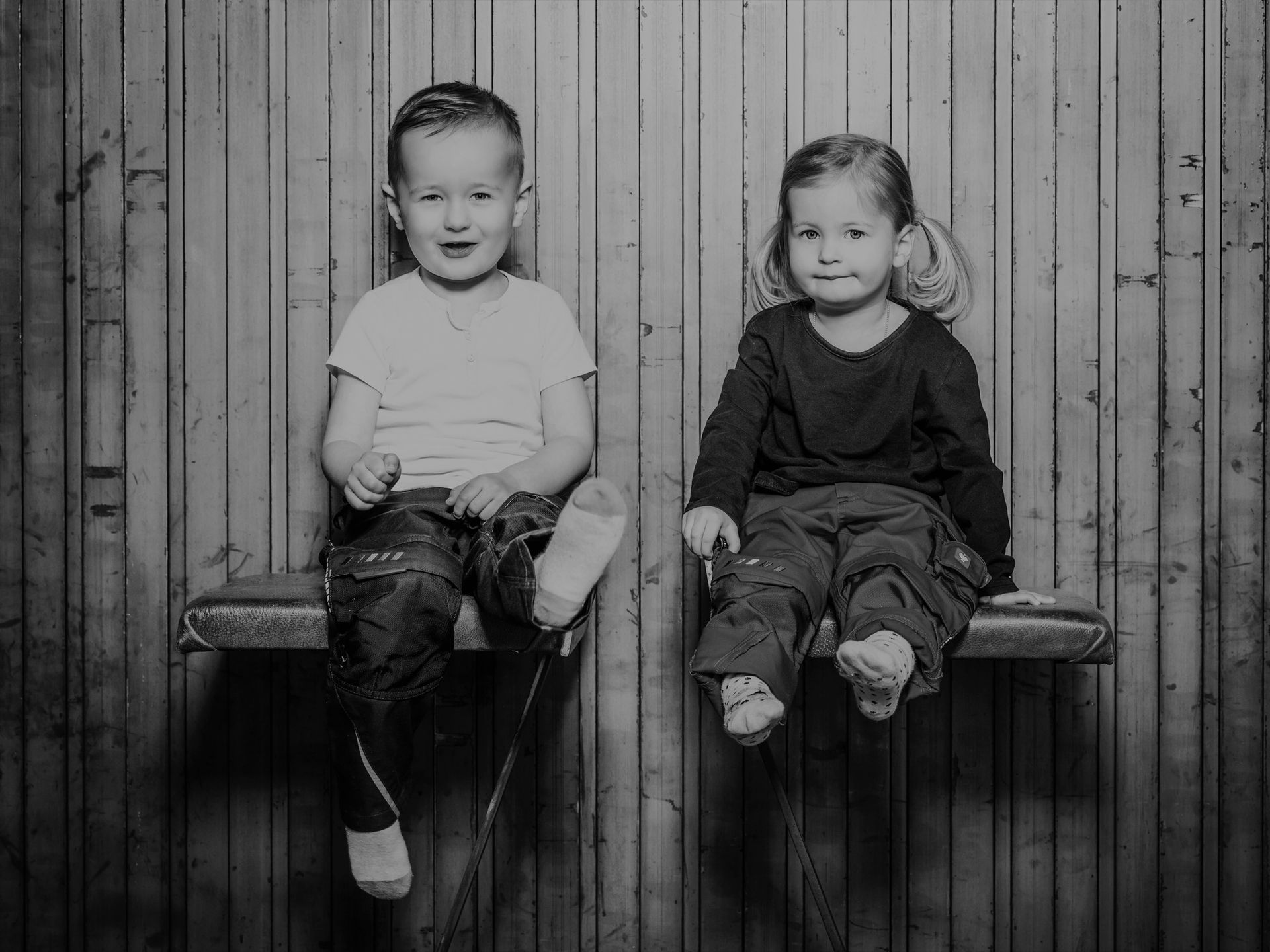 Zwei kleine Kinder sitzen nebeneinander auf einer Bank; der Junge lächelt, das Mädchen schaut nach vorne, vor einem Holzpaneel-Hintergrund.