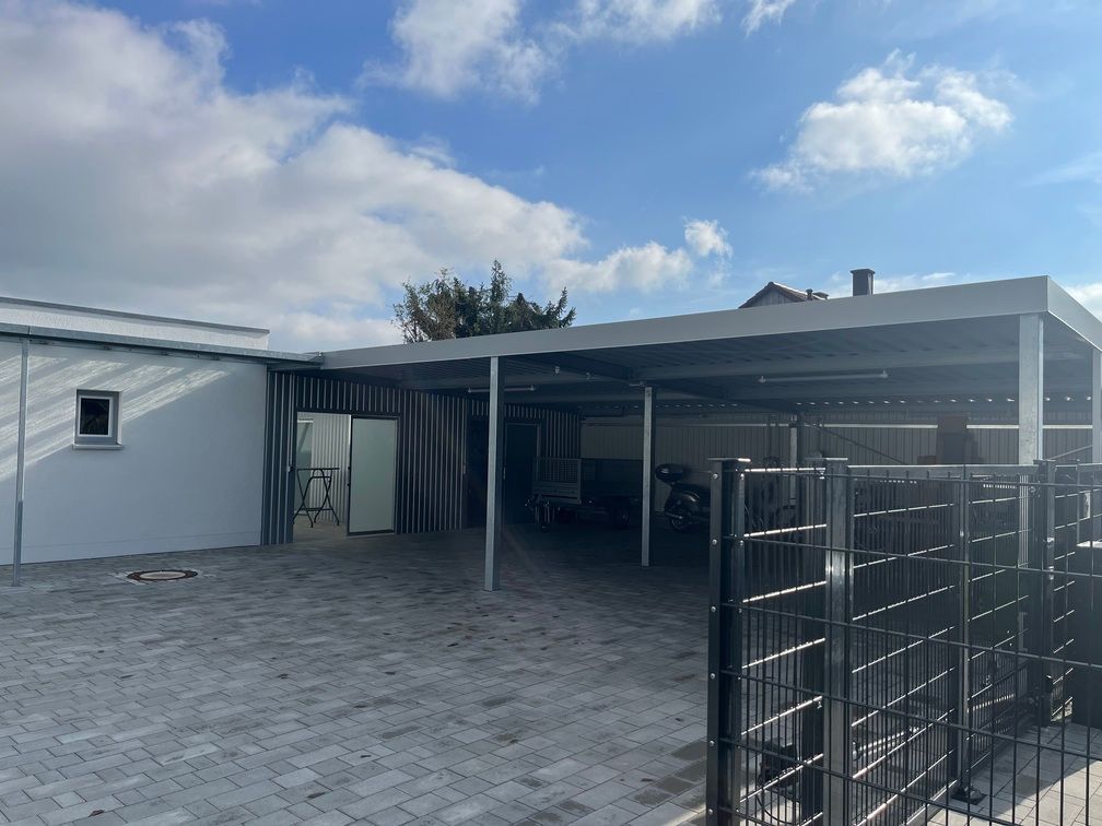 Ein grauer Carport neben einem weißen Gebäude und einem schwarzen Zaun, auf einer grauen Ziegelauffahrt, unter einem bewölkten Himmel.