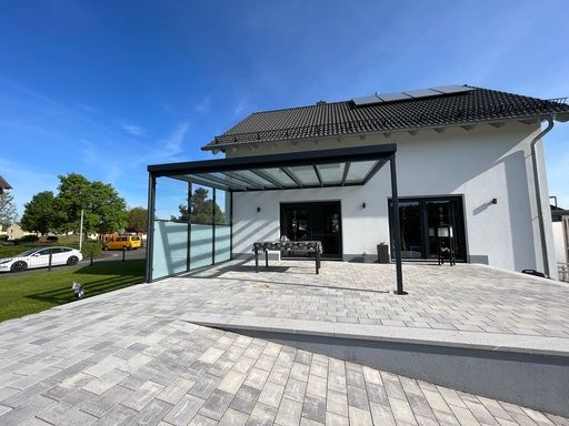 Modernes Haus mit Terrasse, Glasdach, grauen Pflastersteinen und Solarzellen auf dem Dach unter blauem Himmel.