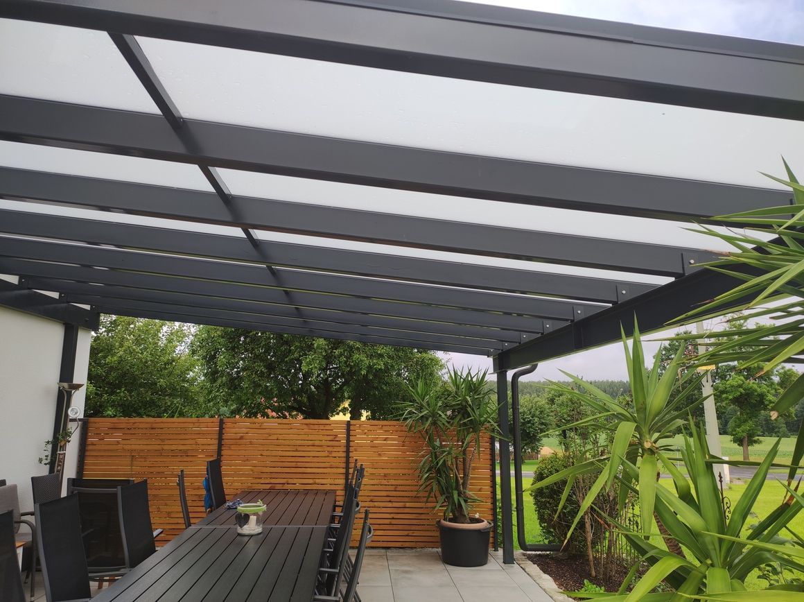 Überdachte Terrasse mit dunkelgrauem Rahmen, durchsichtigen Dachplatten, langem Tisch und Grünpflanzen.