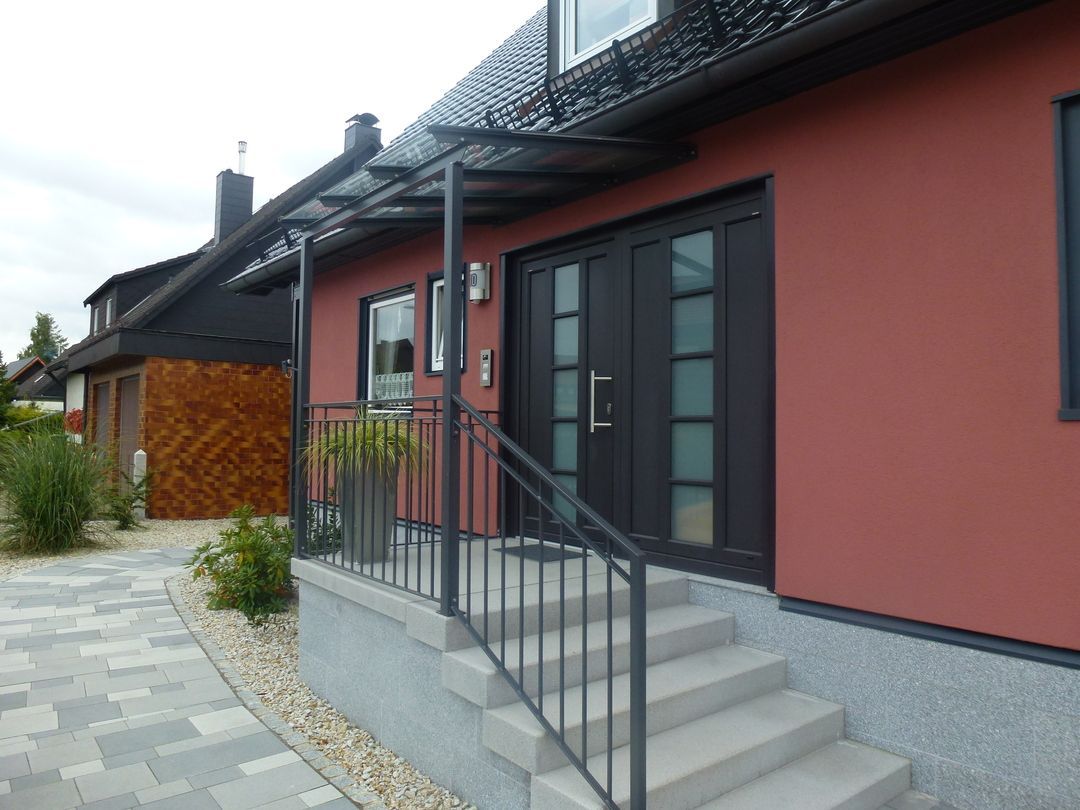Rotes Haus mit dunklen Türen, Veranda und Stufen. Metallgeländer und Glasdach über dem Eingang.
