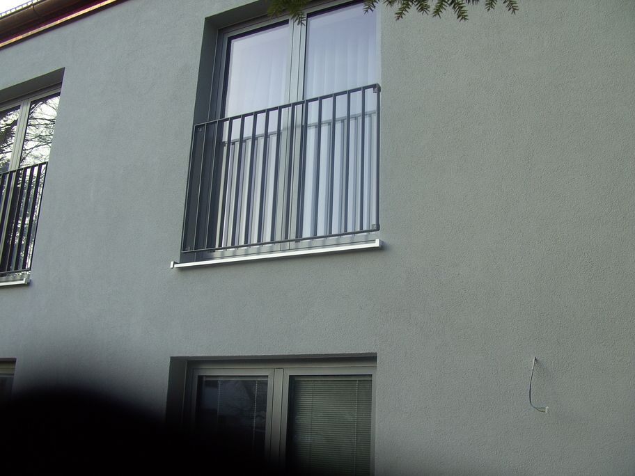 Graues Gebäude mit Balkonen und Metallgeländern; Fenster und Jalousien sichtbar.