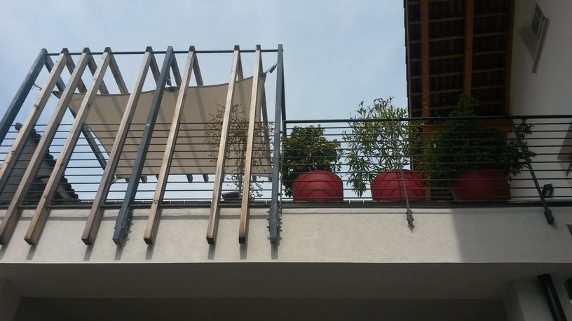 Balkon mit Pflanzen in roten Töpfen, Metallgeländer, Holzlatten und Leinwandschirm unter bewölktem Himmel.