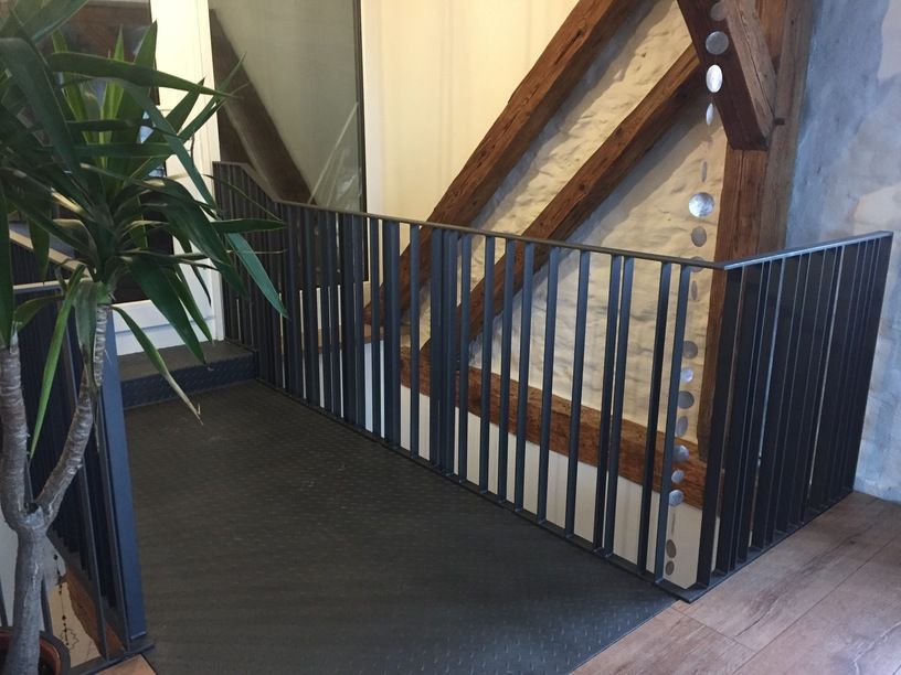 Innenansicht: Schwarzes Metallgeländer auf einem Treppenabsatz vor einer Steinmauer mit freiliegenden Holzbalken.