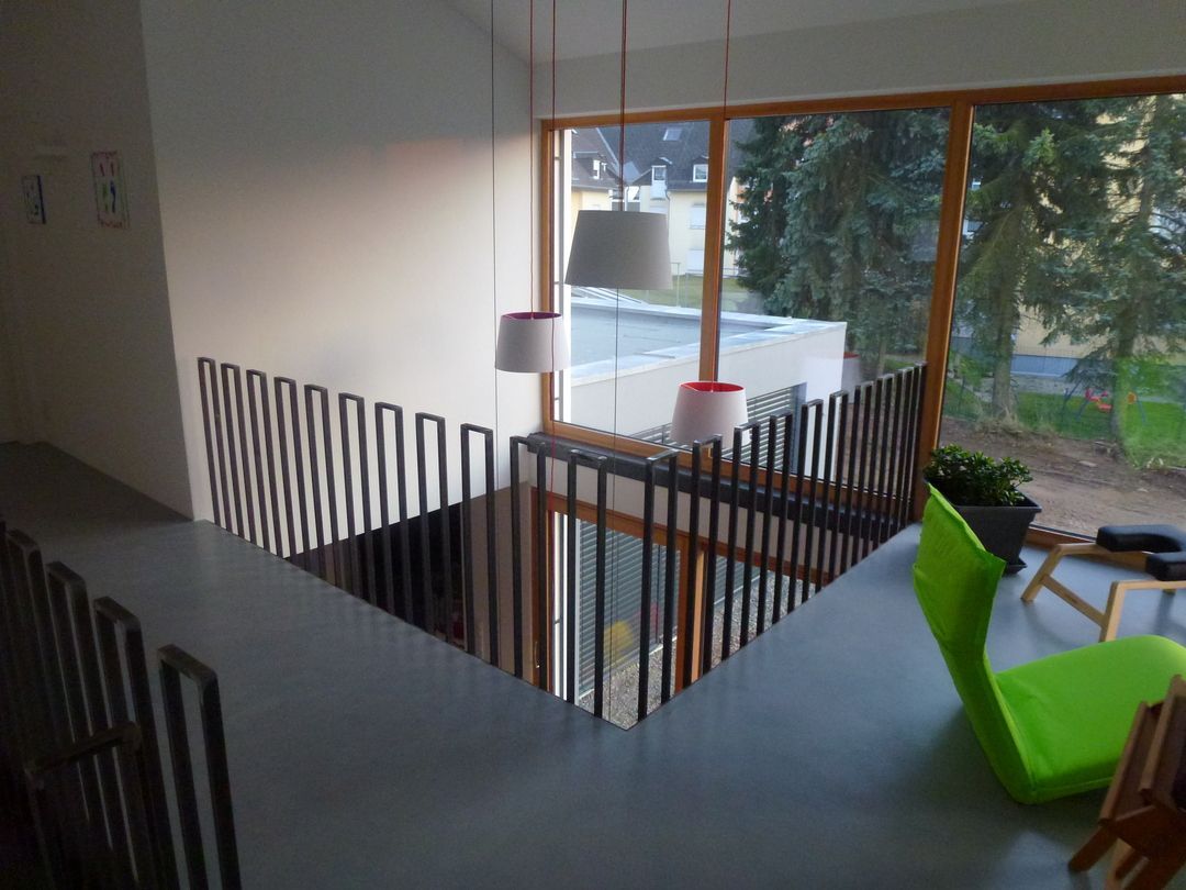 Innenansicht eines modernen Hauses. Ein Metallgeländer umgibt eine Treppe, durch die ein großes Fenster und ein grüner Stuhl sichtbar sind.