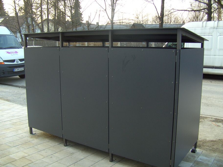 Dunkelgraue Mülltonnenumhausung aus Metall mit Flachdach, neben einem Gehweg und einer Straße.