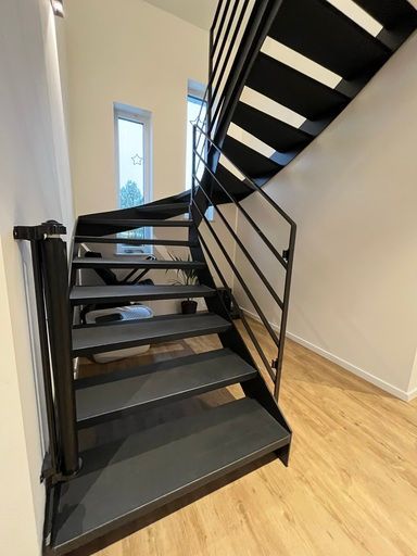 Schwarze Metalltreppe mit horizontalem Geländer, dunkelgrauen Stufen und weißem Wandhintergrund.