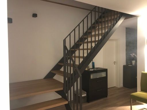 Treppe mit dunklem Metallgeländer und Holzstufen; führt von einem Wohnbereich nach oben.