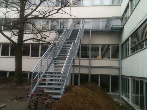 Metallische Außentreppe vor einem Gebäude mit Fenstern und einem Baum.