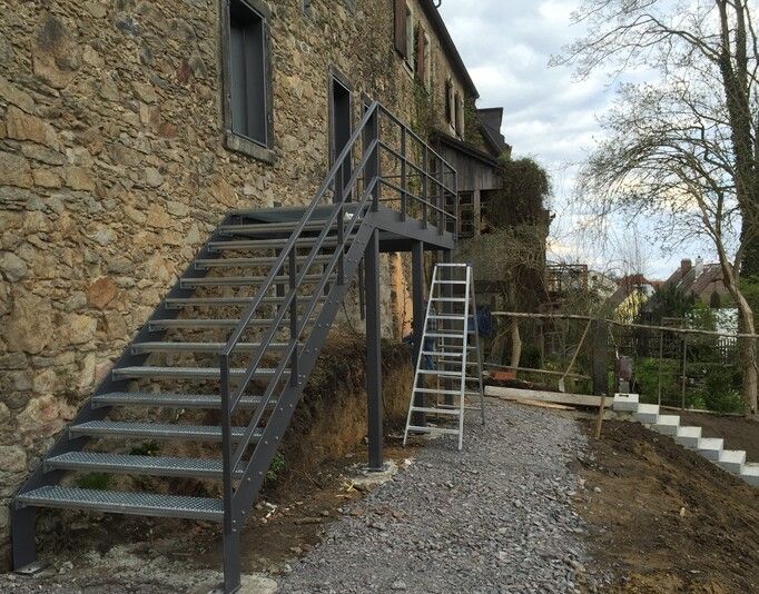 Eine Stahltreppe führt zu einem Steingebäude hinauf, daneben ein Kiesweg und eine Leiter.