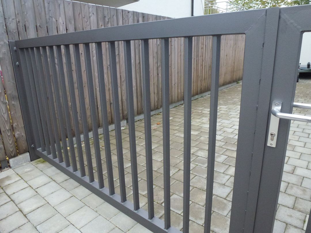 Graues Metalltor mit vertikalen Stäben in einem gepflasterten Bereich, im Hintergrund ein Holzzaun.