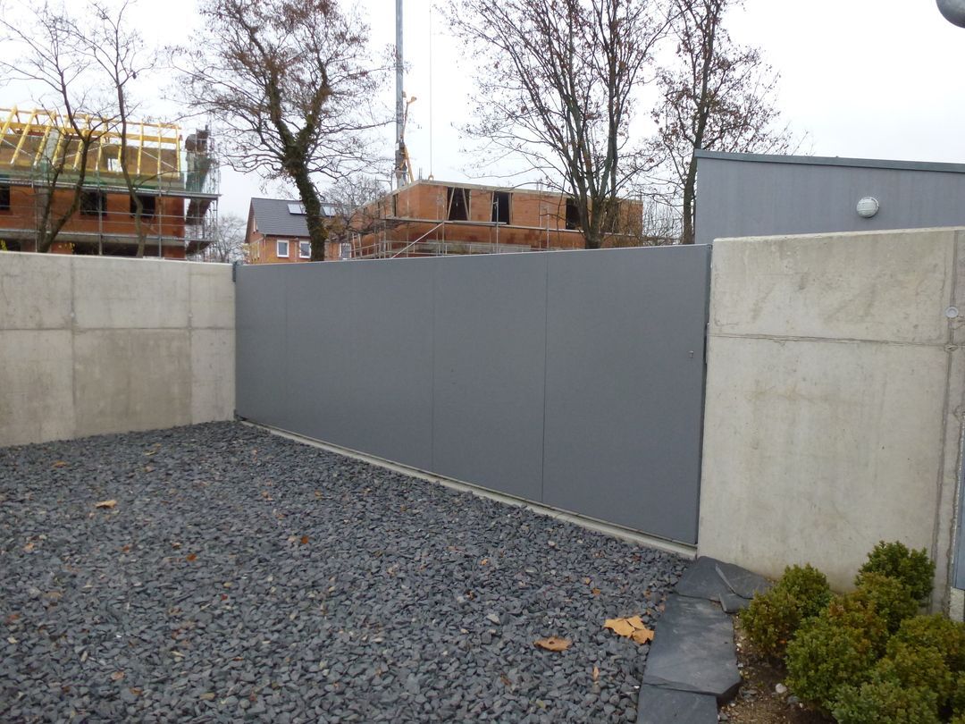 Graues Tor zwischen Betonmauern auf einer Kiesauffahrt. Im Hintergrund Bauarbeiten.