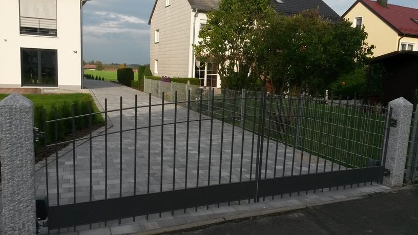 Metalltor und Auffahrt zu einem Haus, graue Pflastersteine, grüner Rasen, Granitpfosten.