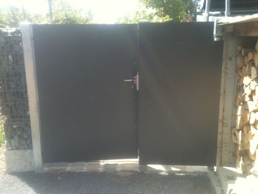 Schwarzes, einbetoniertes Metall-Doppeltor mit Griff; rechts davon Brennholz.