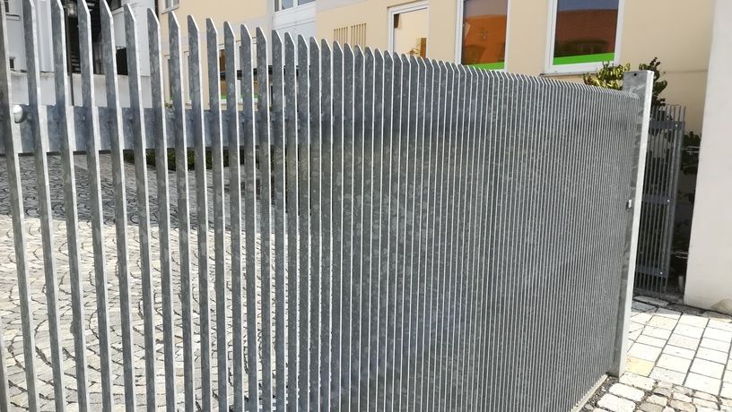 Metallzaun mit vertikalen Stäben und einem geschlossenen Tor vor einem Gebäude.