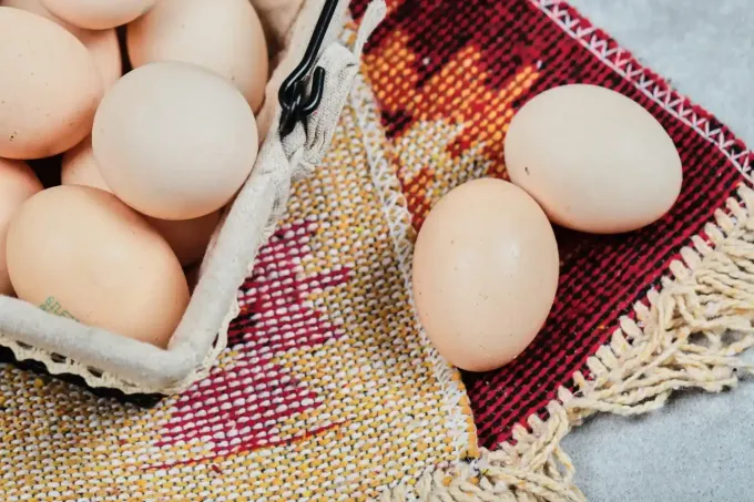 Huevos de color claro reposan en una cesta tejida y sobre una estera de tela estampada con borde de flecos.