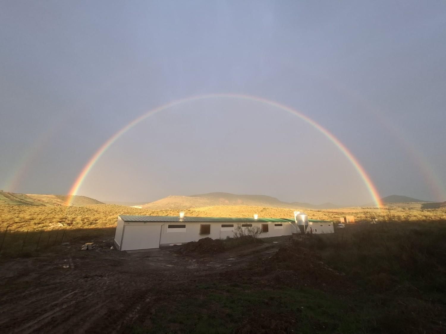 Un doble arcoíris se extiende sobre un edificio largo, blanco y de una sola planta, en un paisaje ondulado.
