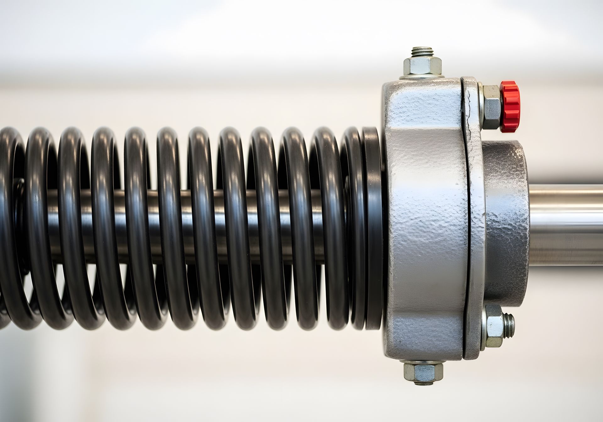Un ressort métallique enroulé entoure une tige d'acier, fixée par un boîtier métallique gris boulonné avec une valve de réglage à capuchon rouge.