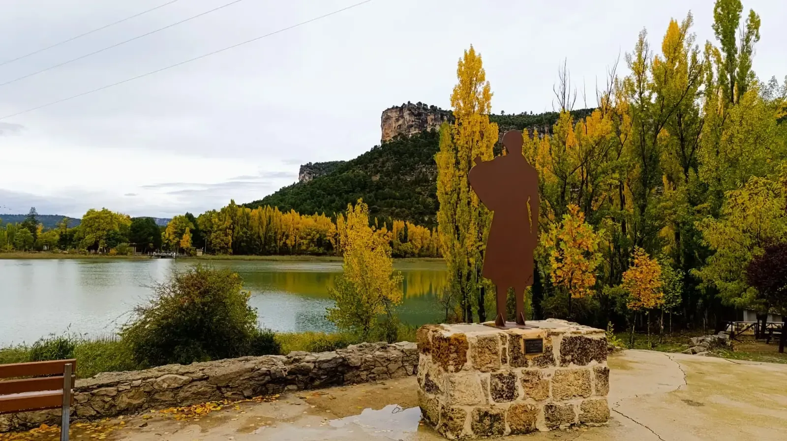 Una estatua de un hombre parado sobre un bloque de piedra frente a un lago rodeado de árboles.