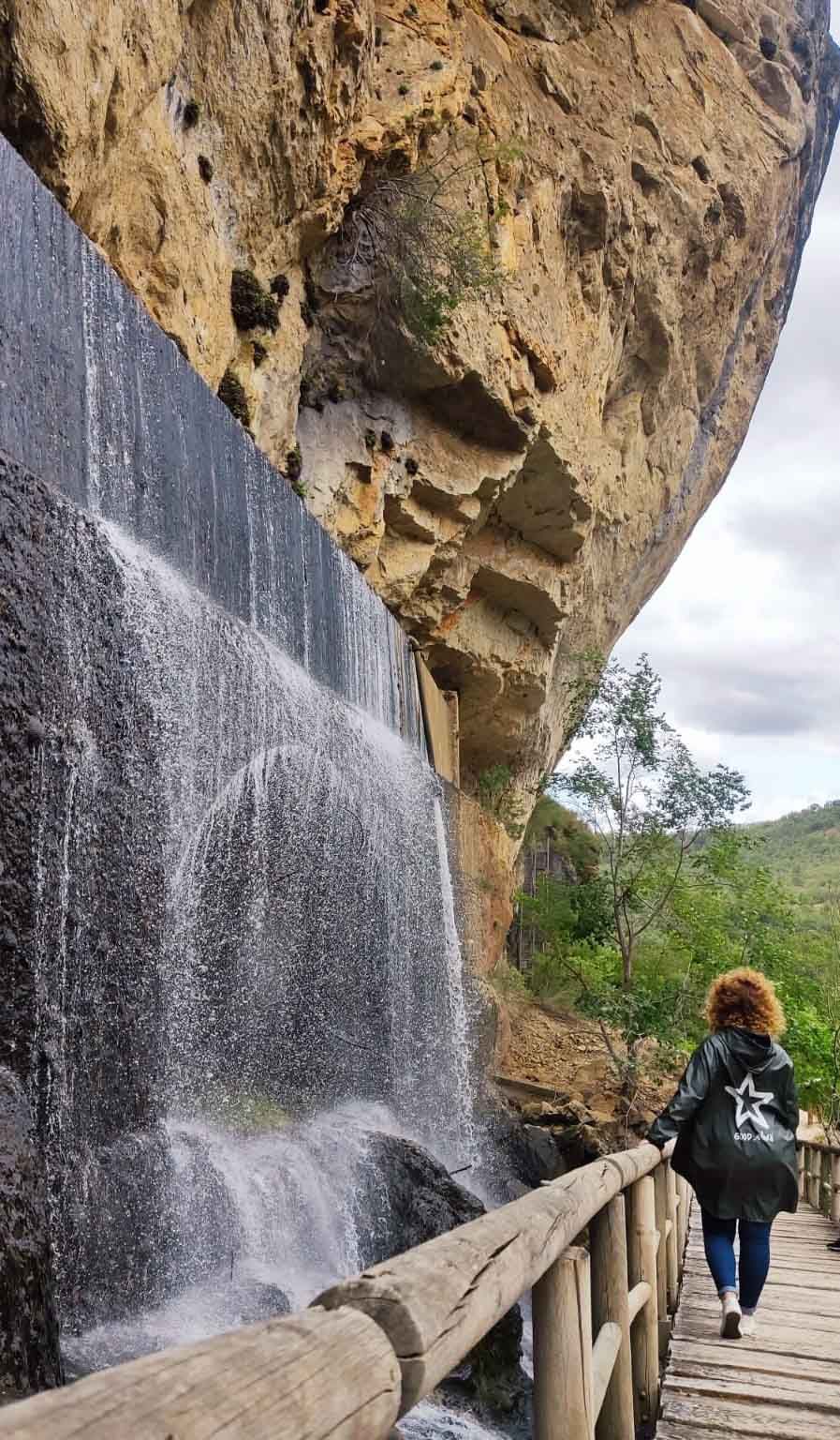 Una mujer camina sobre un puente de madera al lado de una cascada.