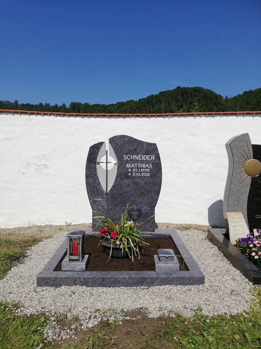 Dunkler Grabstein mit Kreuzmotiv, davor Blumenschmuck und eine rote Grabkerze, umgeben von Kiesfläche vor einer weißen Mauer.