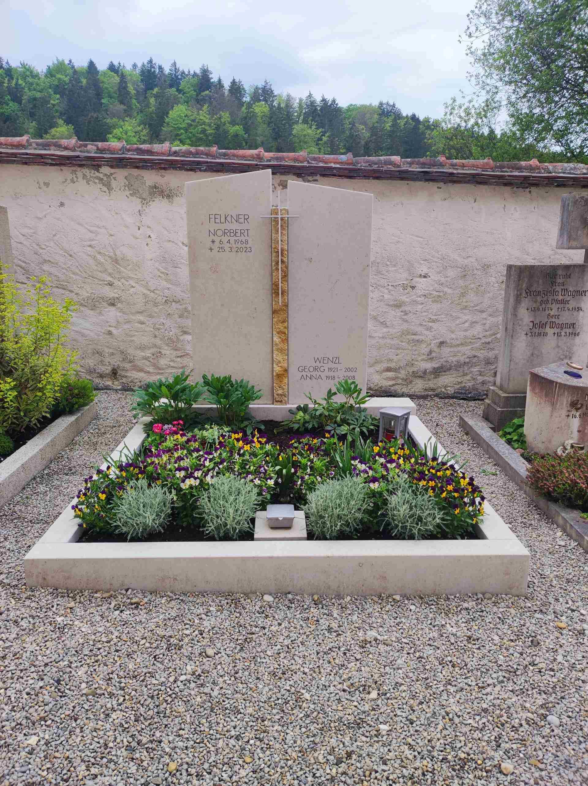 Heller, zweiteiliger Grabstein mit goldfarbener Zierleiste in der Mitte, davor bunt bepflanztes Blumenbeet und zwei Laternen, umgeben von Kiesfläche.