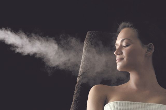 Frau mit geschlossenen Augen geniesst den Dampf vor dunklem Hintergrund.