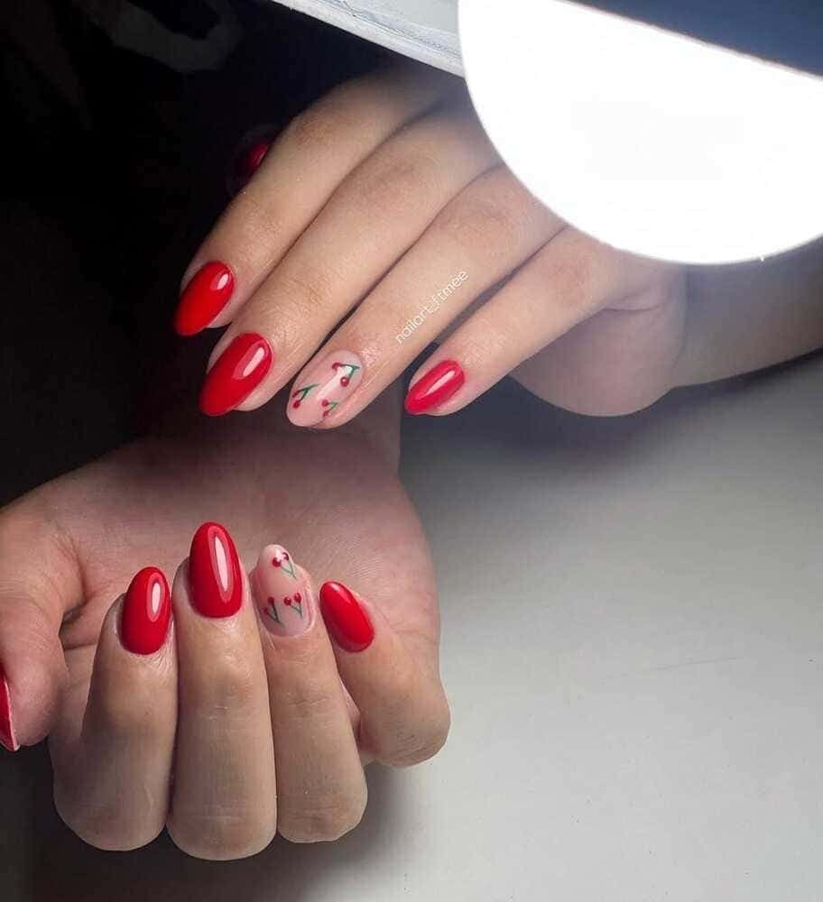 Manos con uñas rojas en forma de almendra, una uña decorativa con diseño de cereza, colocada sobre una superficie blanca.