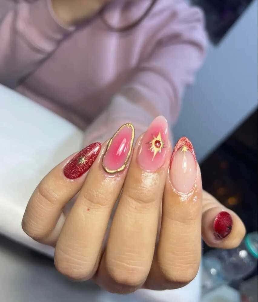 Mano con elegantes uñas almendradas. Brillantina roja, degradados rosas y detalles dorados crean un diseño moderno.
