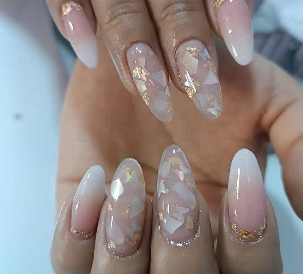 Uñas de forma ovalada con un degradado rosa-blanco, adornadas con copos dorados y piezas iridiscentes.