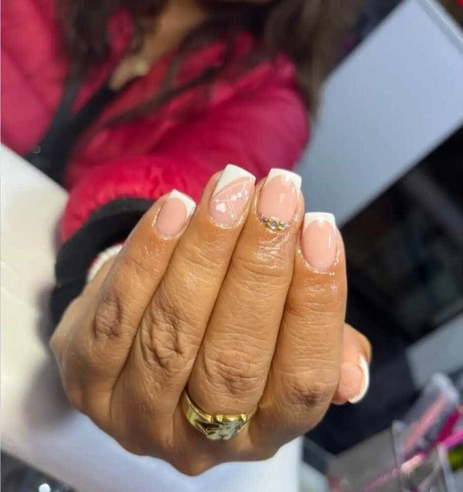 Mano con manicura francesa, puntas blancas y anillo dorado. Chaqueta rosa al fondo.