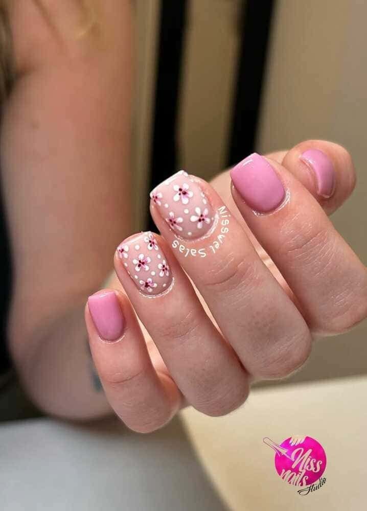 Uñas rosas mate, dos con diseño floral blanco, mano delante de fondo borroso.