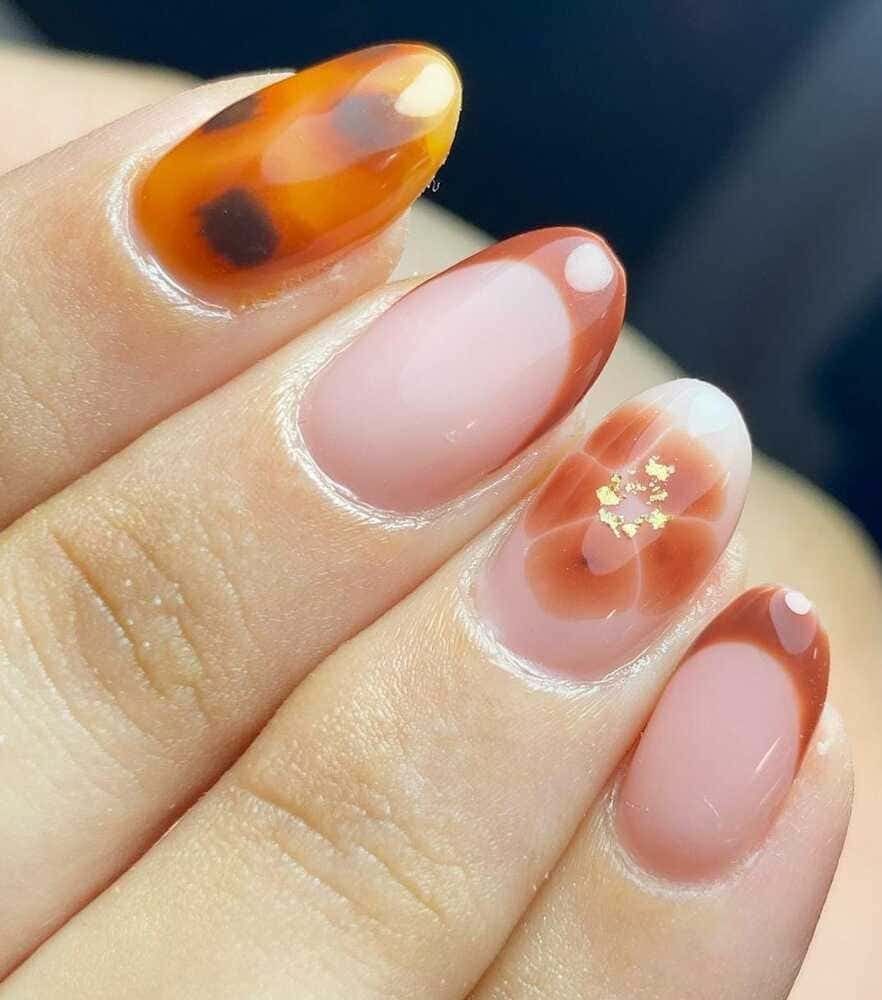 Primer plano de una mano con uñas de forma ovalada, con diseños de carey, florales y de punta francesa en marrón, blanco y rosa.