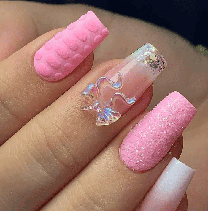 Uñas acrílicas rosas con detalles texturizados y brillantes, además de un acento de lazo 3D.