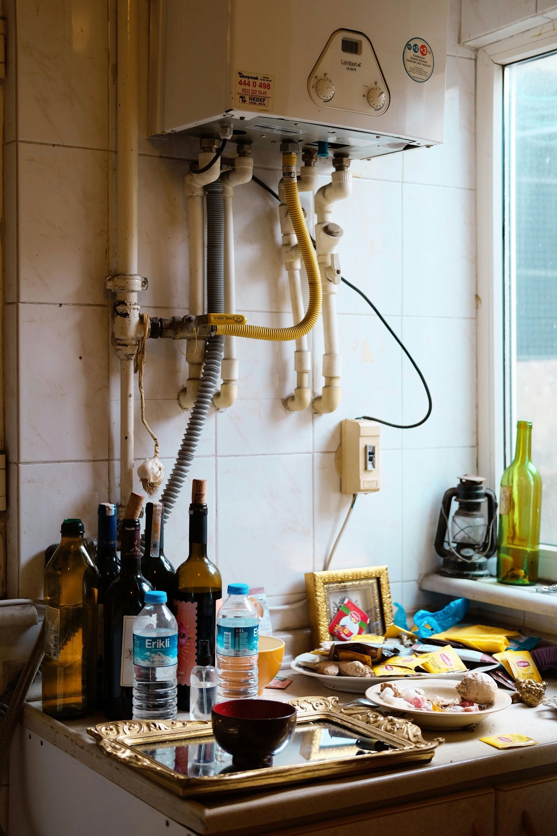 Mostrador de cocina con botellas de vino, comida y un calentador de agua contra una pared de azulejos blancos.