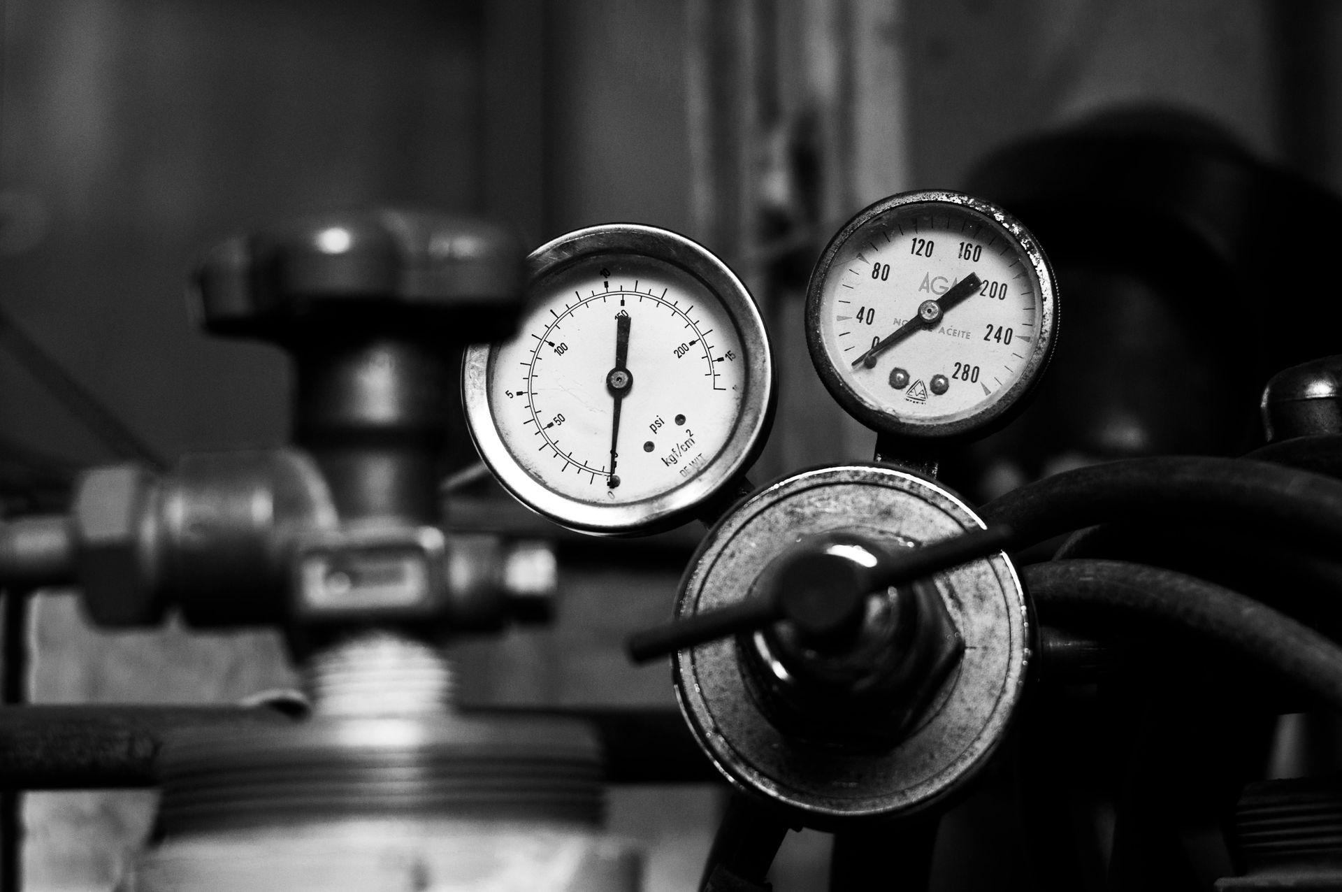 Primer plano en blanco y negro de medidores en equipos industriales; se ven válvulas, tubos de metal y diales.