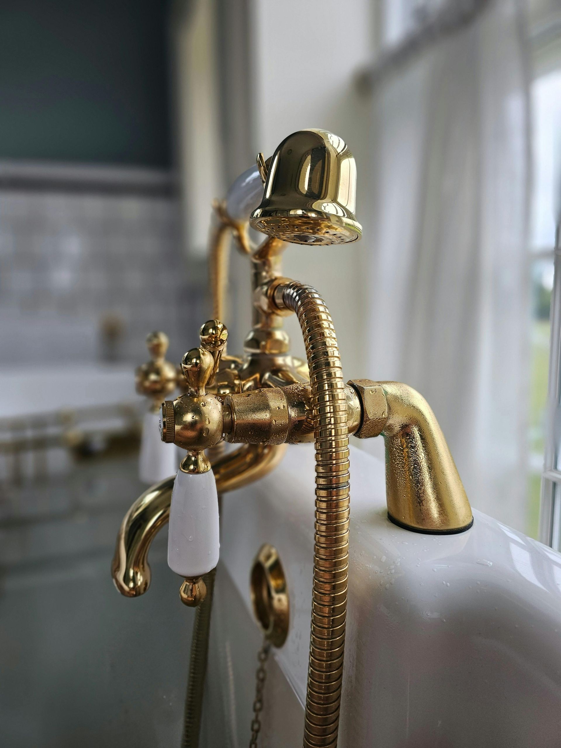 Grifo de baño bañado en oro con manijas blancas, sobre una bañera blanca, cerca de una ventana con cortinas transparentes.