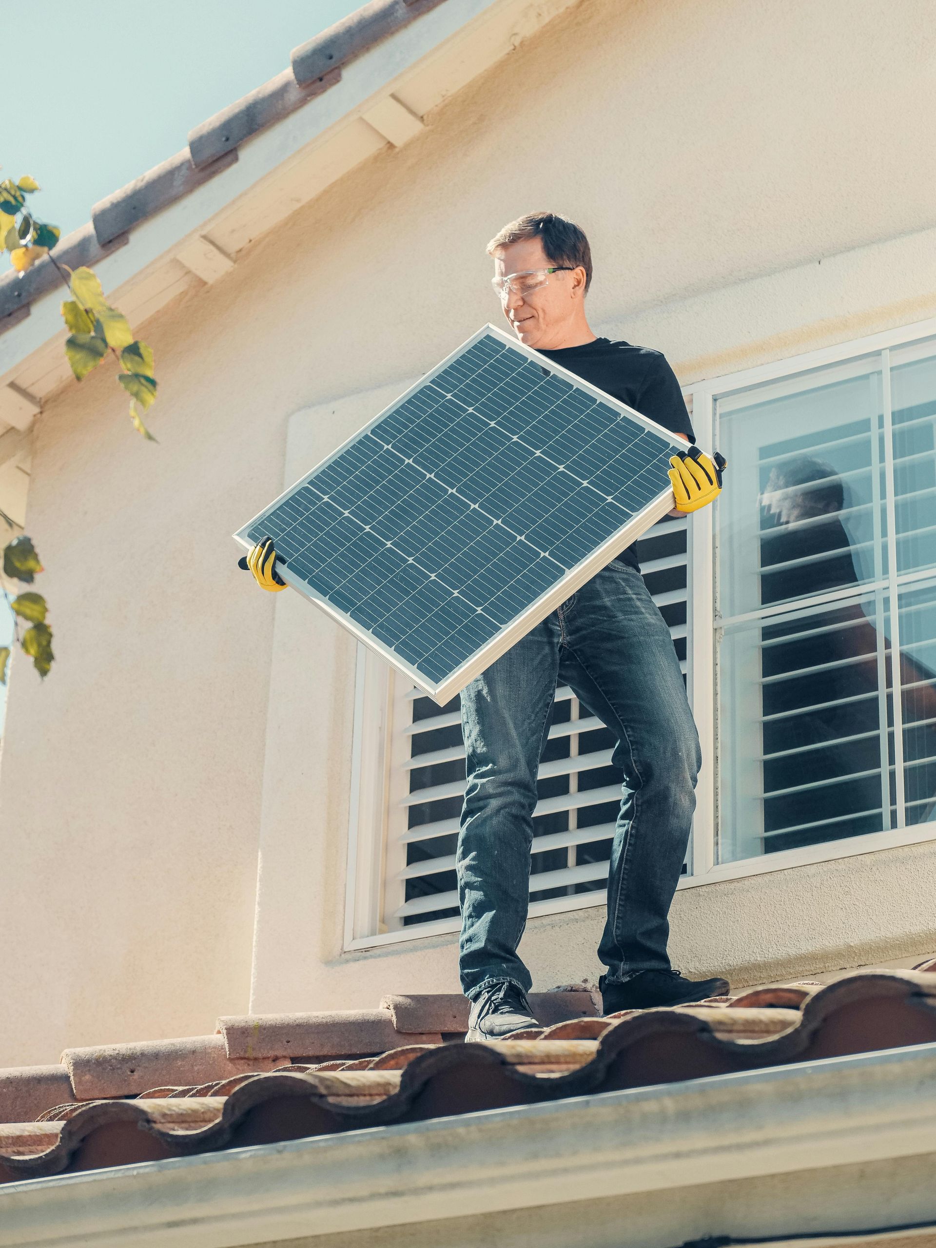 Un hombre lleva un panel solar en un tejado, con guantes. Día soleado, casa beige, ventana al fondo.