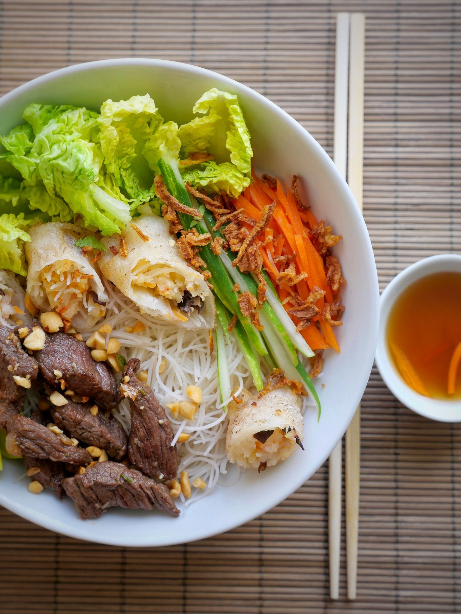 Bol de nouilles vermicelles vietnamiennes avec viande, rouleaux de printemps et légumes, servi avec sauce et baguettes.