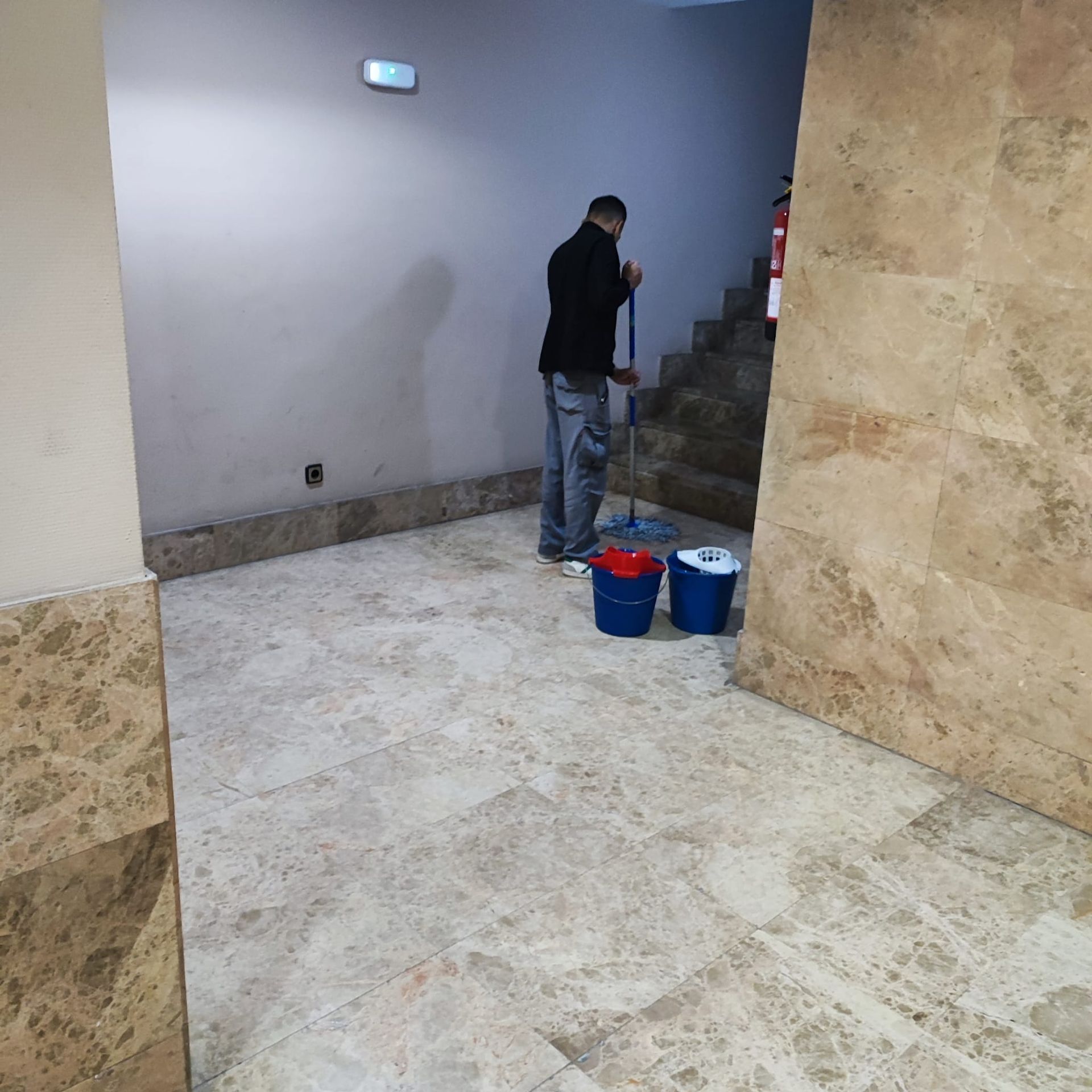 Un hombre fregando un suelo de baldosas en un pasillo. Se ven cubos y escaleras.