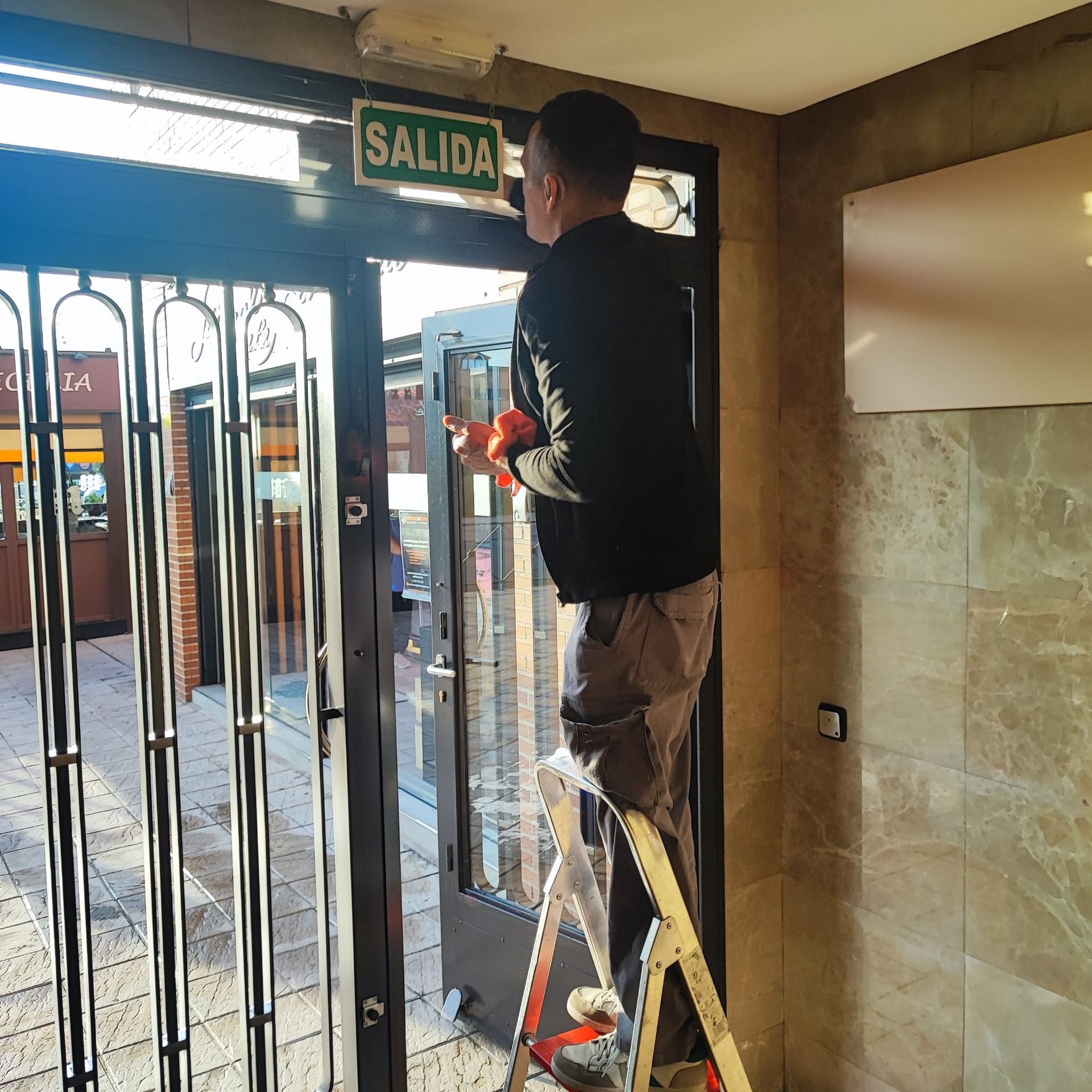 Un hombre en una escalera ajusta un cartel de salida de 'SALIDA' encima de una puerta de vidrio con barras de seguridad de metal.